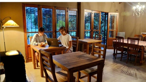 Un'area pranzo con tavoli e sedie in legno, ampie finestre vista alberi e un lampadario.