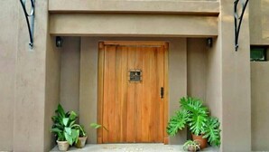 Una porta di legno con una placca di metallo, affiancata da piante in vaso, che conduce a un edificio con facciata in cemento e ingresso piastrellato.