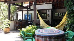 Un patio exterior con una pérgola de madera, una hamaca, una mesa redonda con mantel de crochet y sillas de plástico verde.