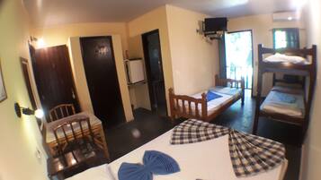 A room with a bunk bed, a single bed, a desk, and a chair. There is a door, a window, and a television mounted on the wall.