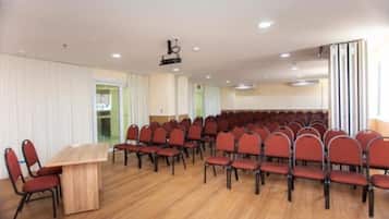 Uma sala de conferência com fileiras de cadeiras vermelhas, uma mesa de madeira e um projetor no teto.