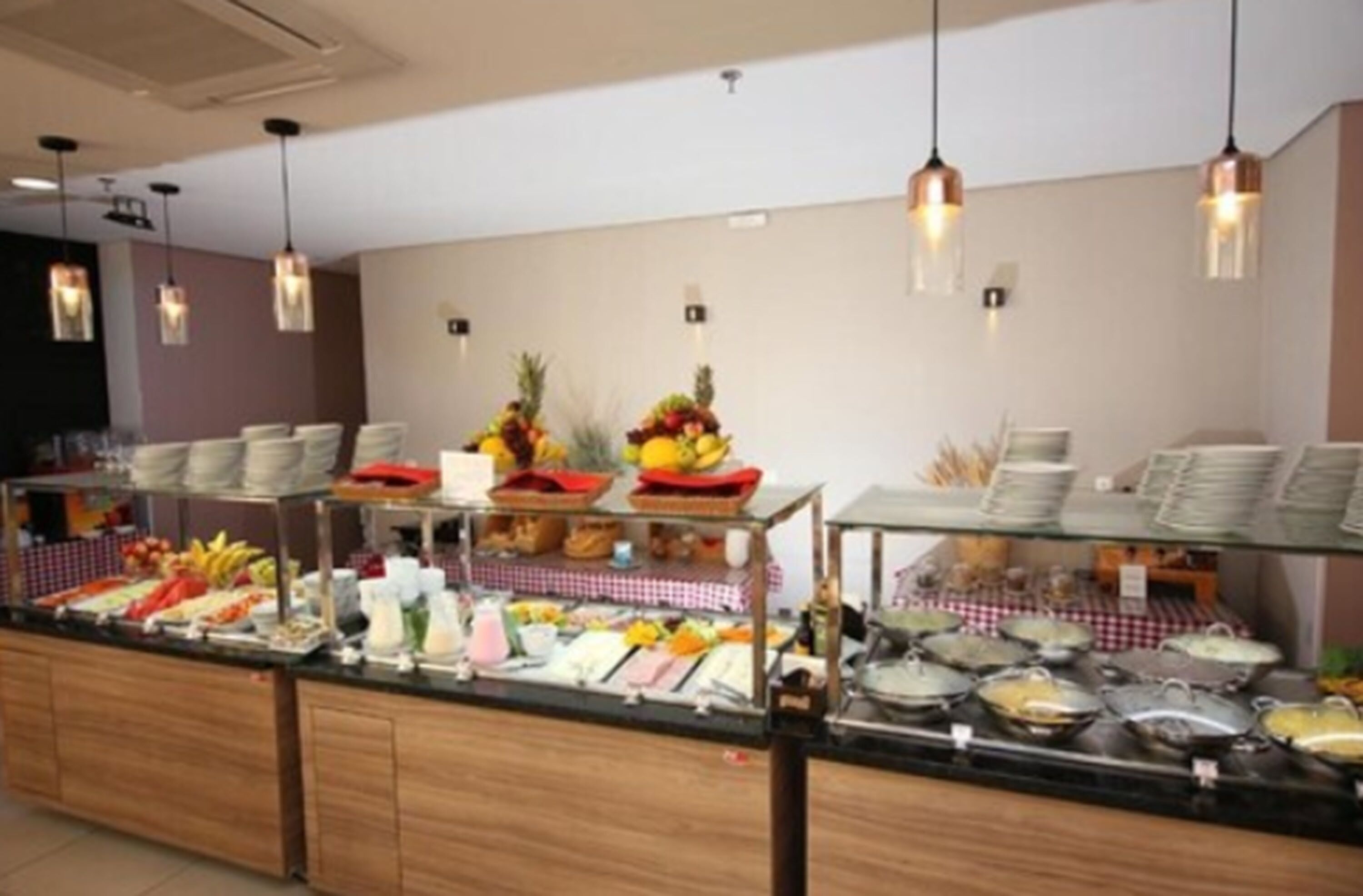 Buffet com diversos alimentos e bebidas, incluindo frutas e pratos, disposto em um balcão com luzes penduradas.