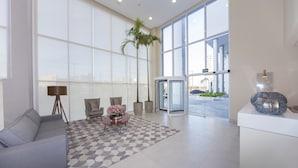 A modern lobby with a grey sofa, a patterned rug, a small table with flowers, a palm tree, and a revolving door. - NB Hotéis (Aracaju)