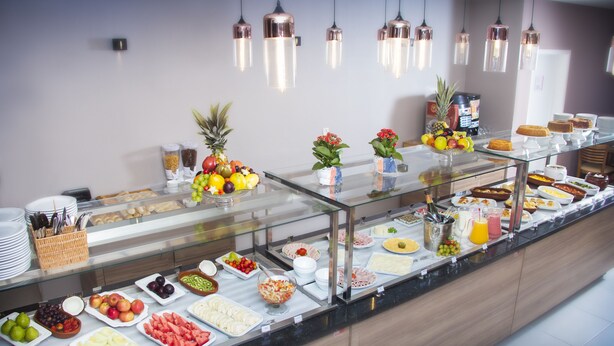 A buffet with various food items and beverages, including fruits, vegetables, and desserts, displayed on a glass counter with hanging lights above. - NB Hotéis (Aracaju)