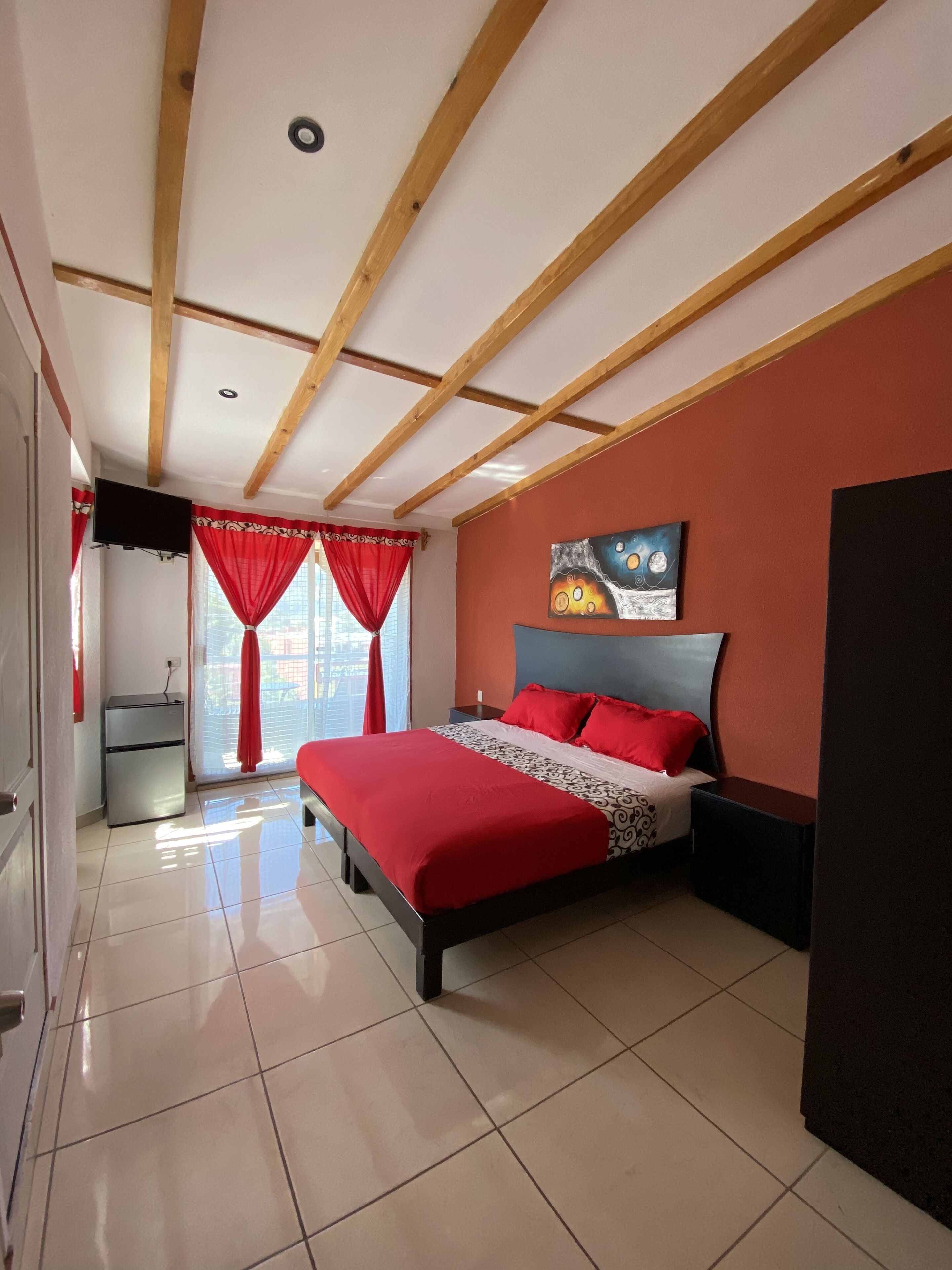 A bedroom with a bed, a TV, a small fridge, and a window with curtains.