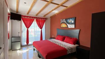 A bedroom with a bed, a TV, a small fridge, and a window with curtains.