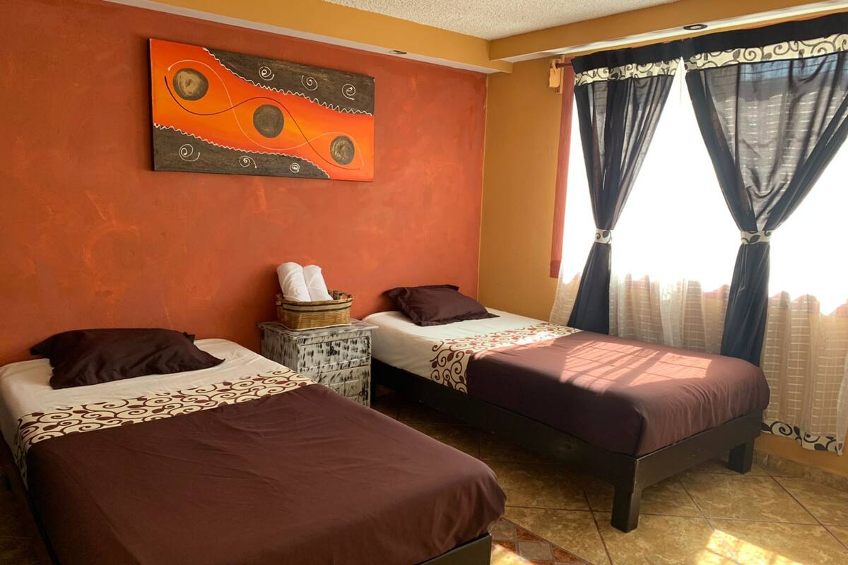 Une chambre avec deux lits, un tableau au mur et des rideaux à la fenêtre.
