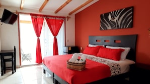 A hotel room with a bed, a TV, a desk, and a window with red curtains.