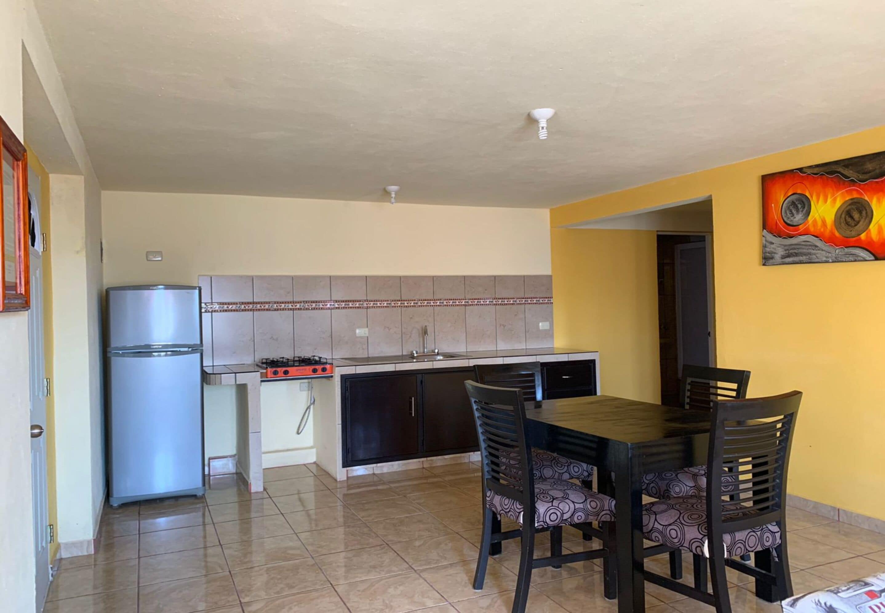 A compact kitchen with a refrigerator, stove, and tiled backsplash. There is a dining table with four chairs.