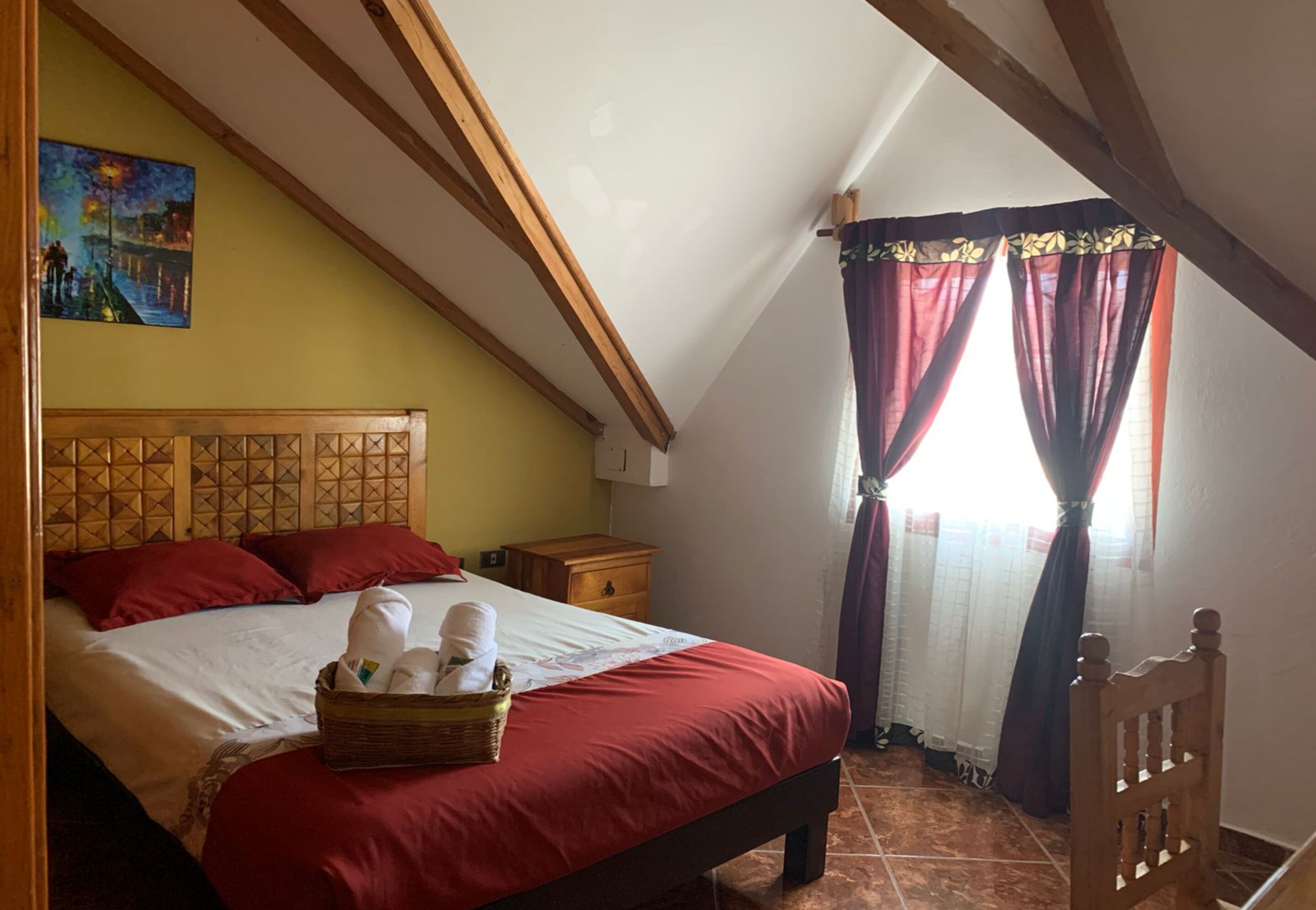 A bedroom with a wooden headboard, a bed with red bedding, a basket of towels, a nightstand, a wooden chair, and a painting on the wall.