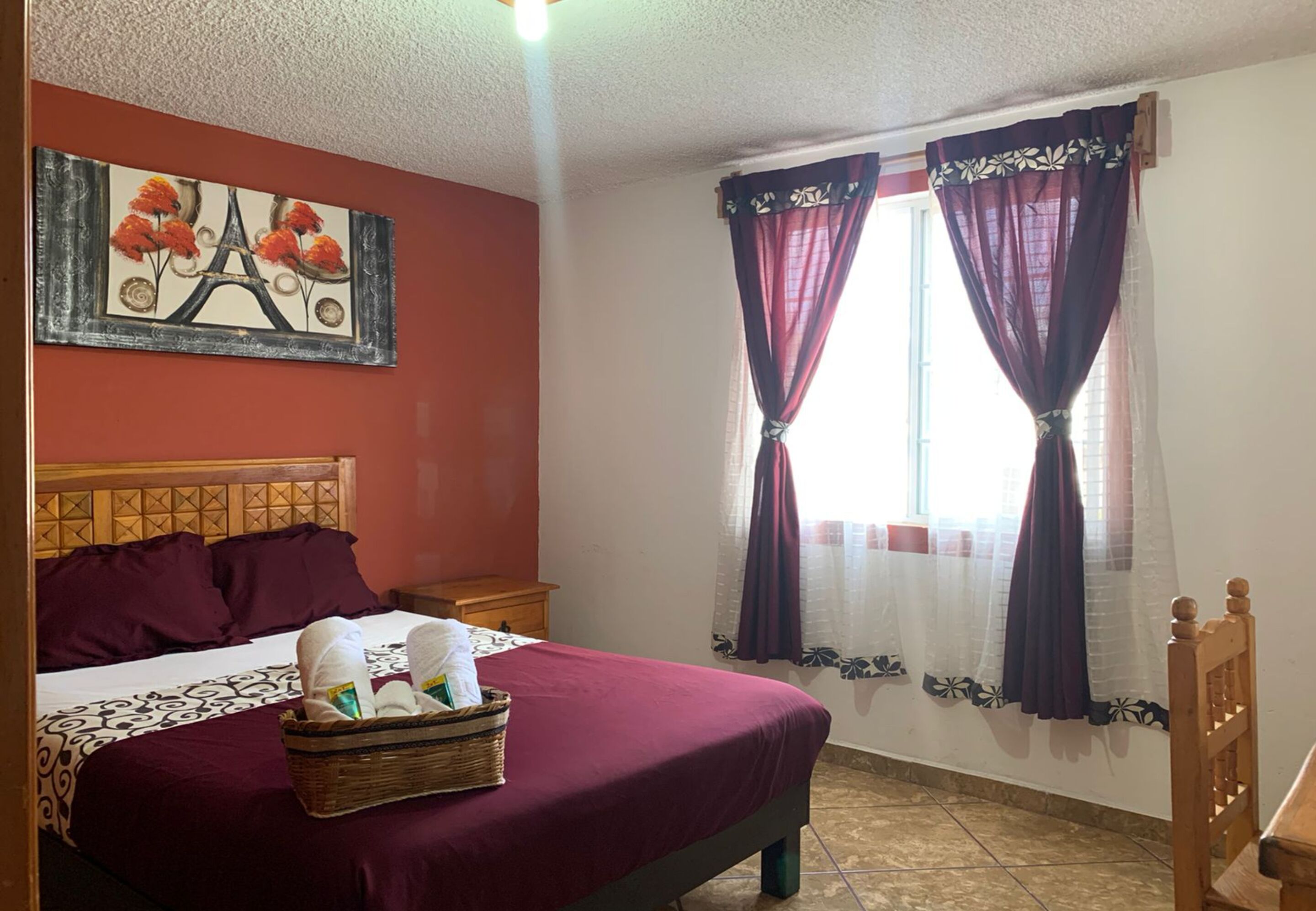 Habitación de hotel con cama, cabecero, mesita de noche, cesta de toallas, un cuadro de la Torre Eiffel y dos ventanas con cortinas.