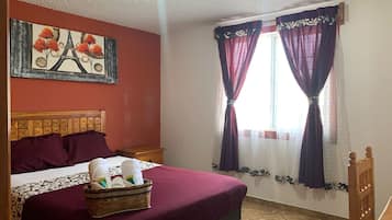 Habitación de hotel con cama, cabecero, mesita de noche, cesta de toallas, un cuadro de la Torre Eiffel y dos ventanas con cortinas.
