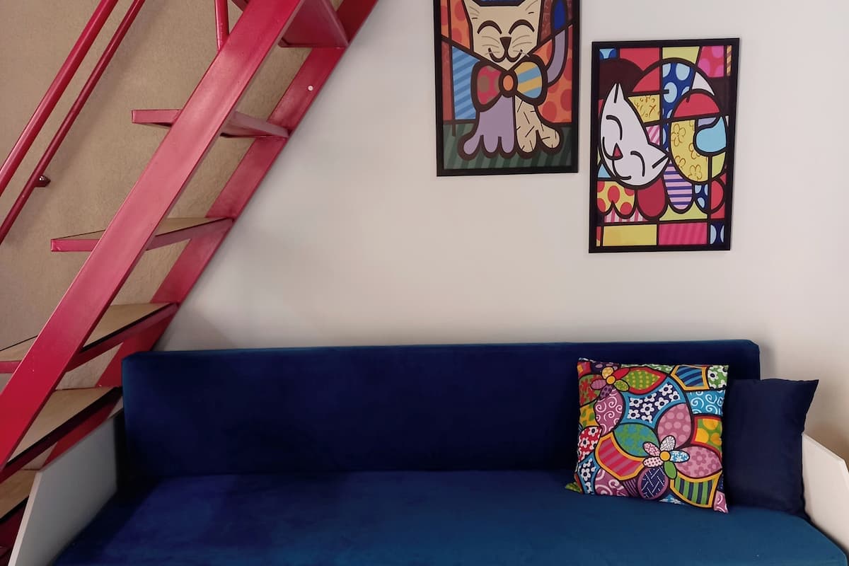 Un canapé bleu agrémenté d’un coussin coloré, un mur blanc et deux œuvres d’art encadrées représentant des chats.