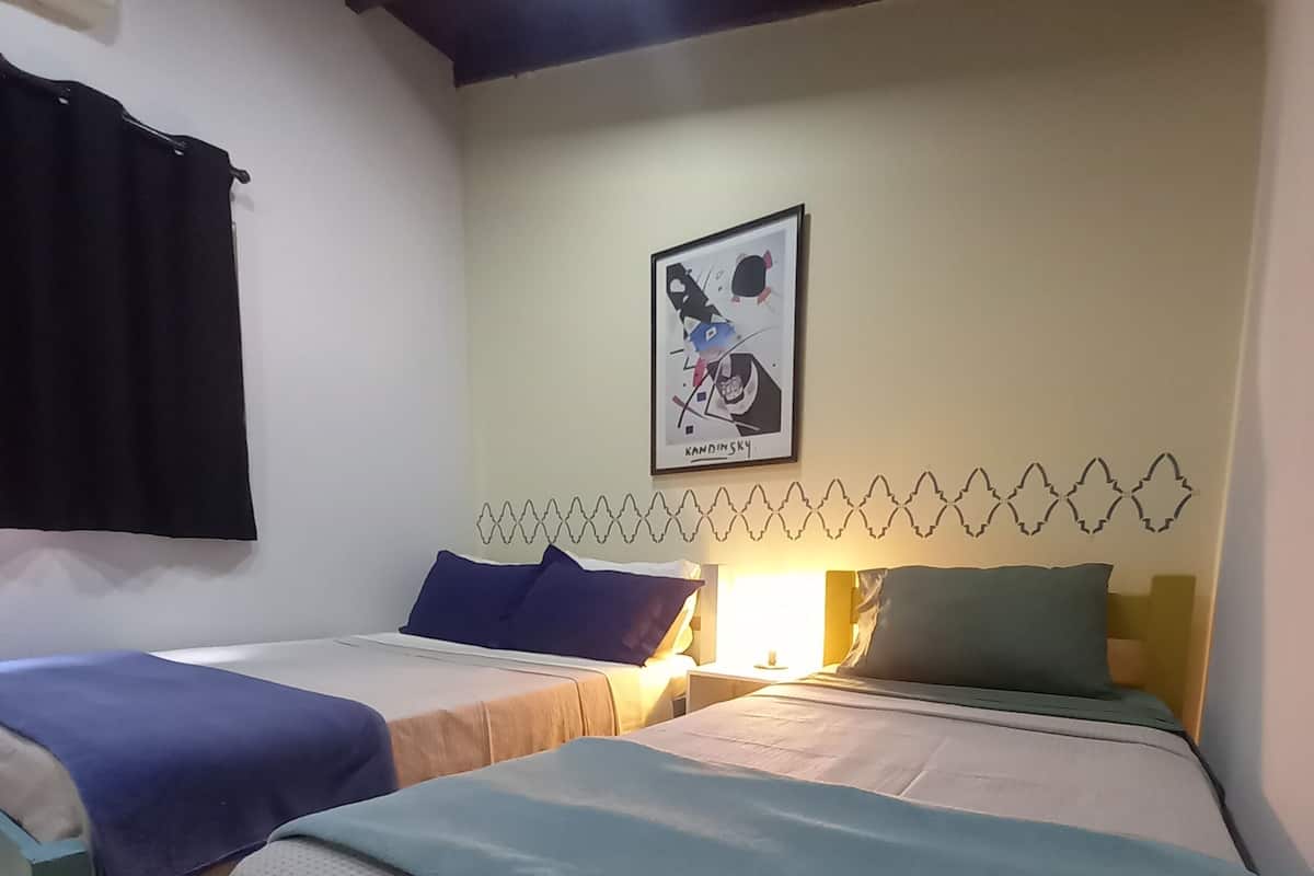 Une chambre avec deux lits, chacun recouvert d’une couverture bleue, un climatiseur fixé au mur et un tableau encadré.