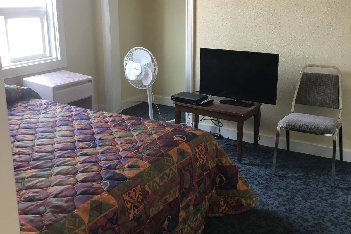 Ett hotellrum med en säng, en tv, en stol, en fläkt och en garderob.