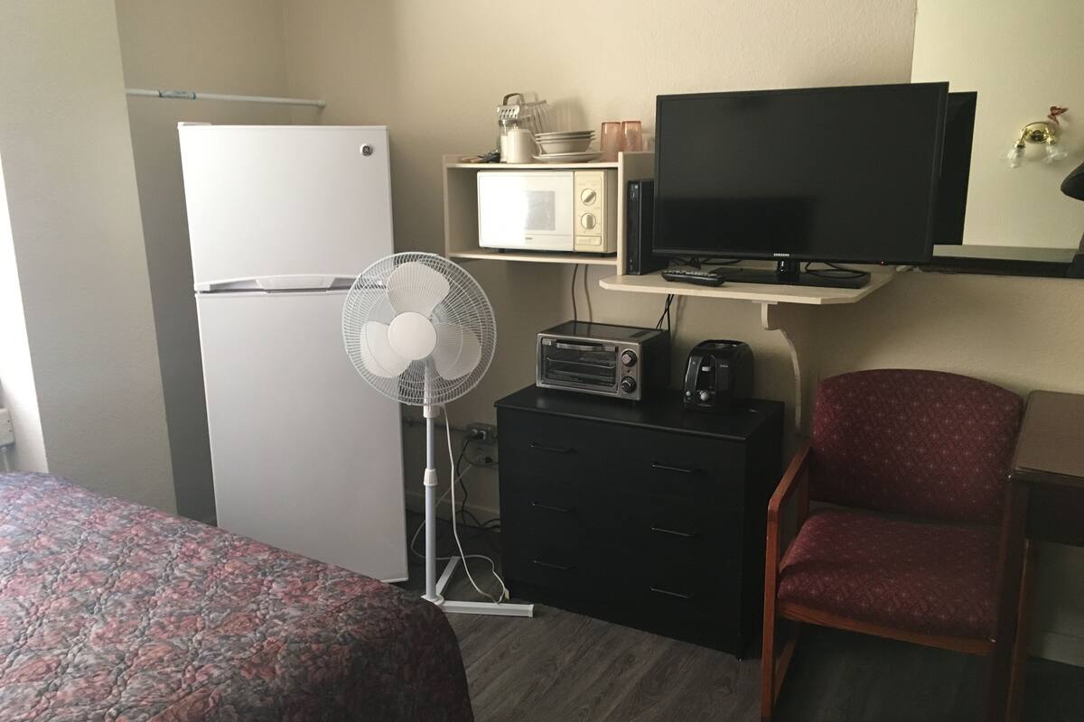 Ett hotellrum med en säng, ett skrivbord med en dator, en stol, en mikrovågsugn, ett kylskåp och en TV.