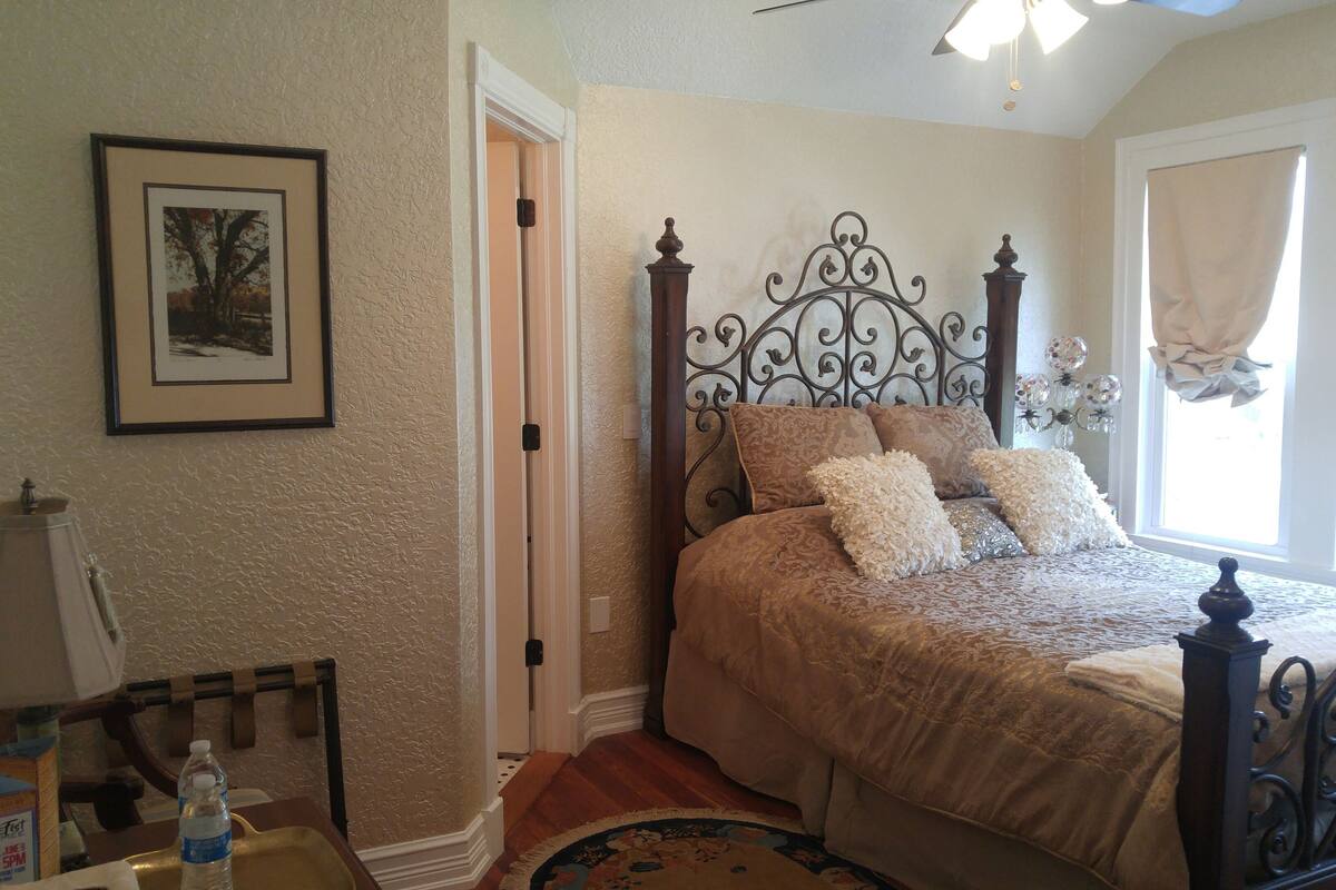 A bedroom with a bed, a nightstand, a lamp, a framed picture, and a window with curtains.