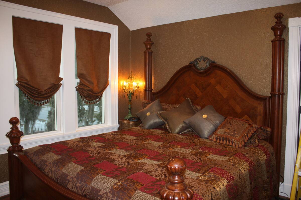 A bedroom with a large bed, a wooden headboard, and two windows with curtains.