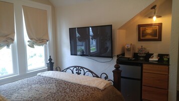 Ein Schlafzimmer mit einem Bett, einem an der Wand befestigten Fernseher, einem Schreibtisch mit Kaffeemaschine und einem Fenster mit Vorhängen.