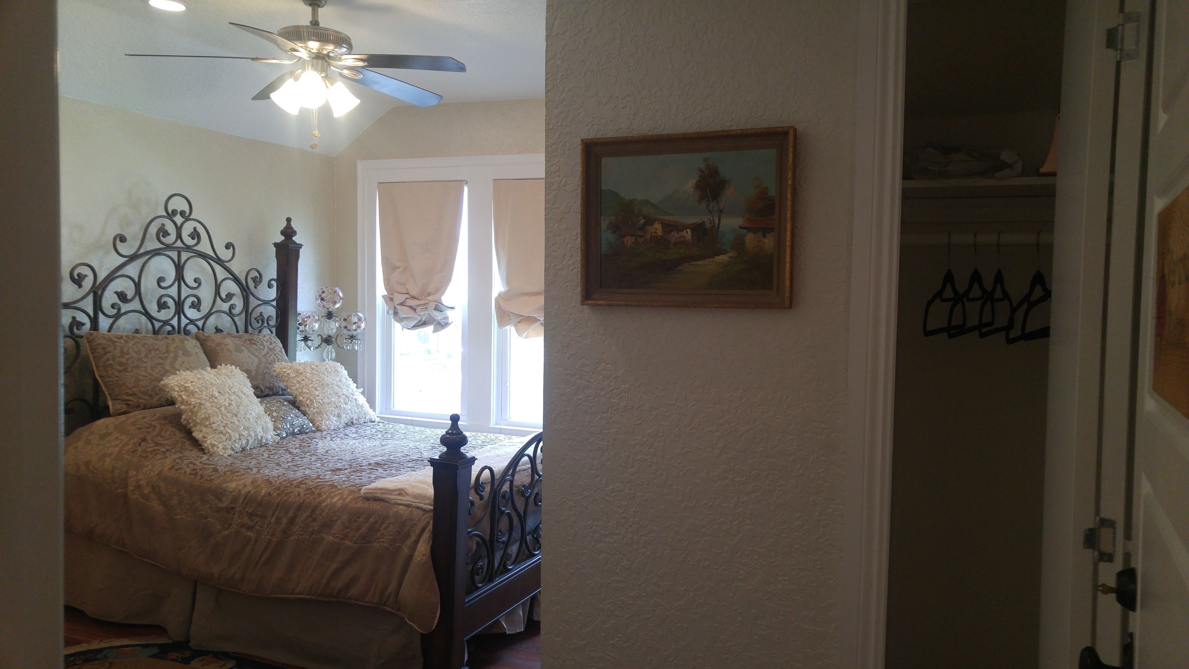 A bedroom with a bed, a ceiling fan, a window with curtains, and a painting on the wall.