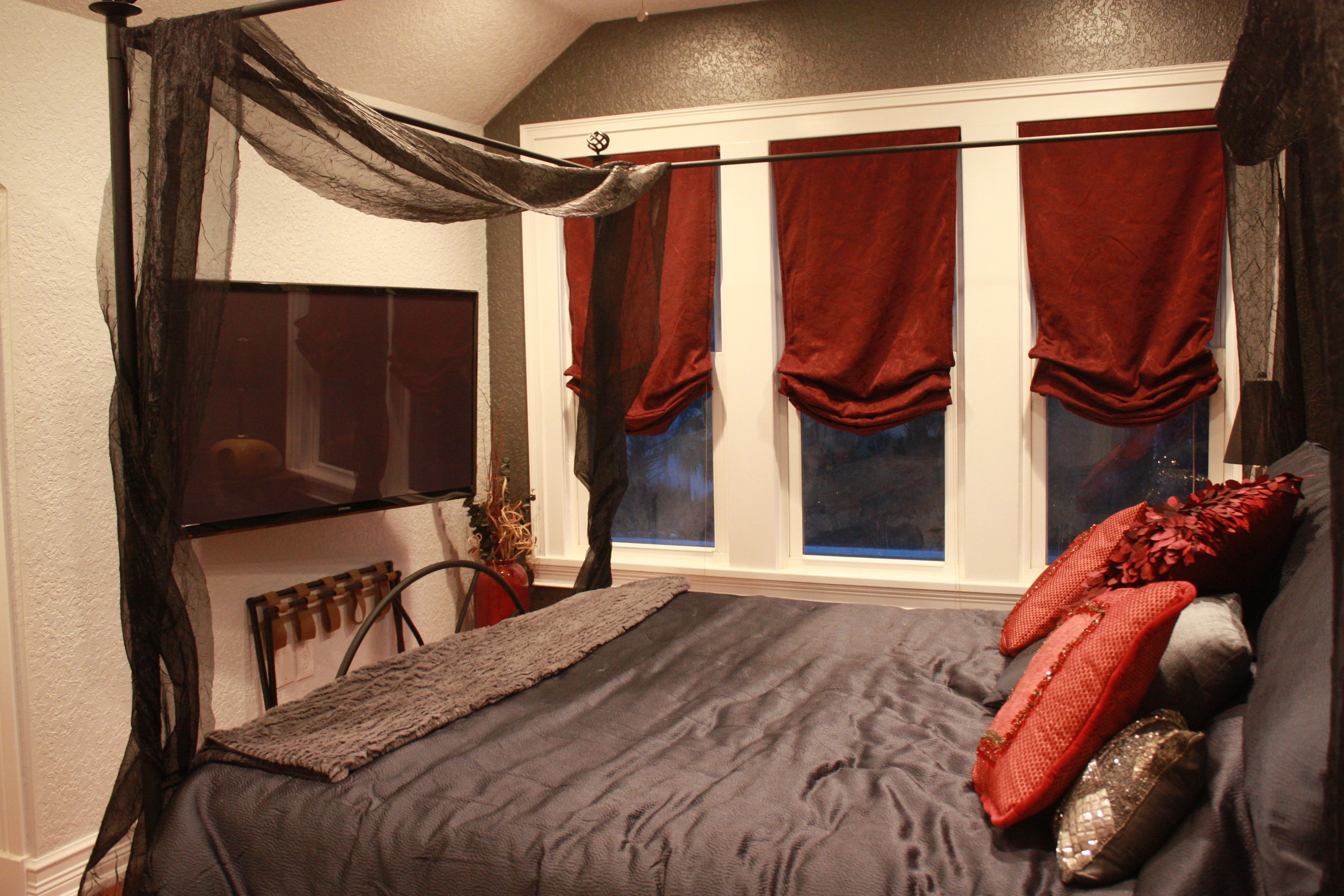 Un dormitorio con una cama grande, un televisor montado en la pared y una ventana con cortinas rojas.