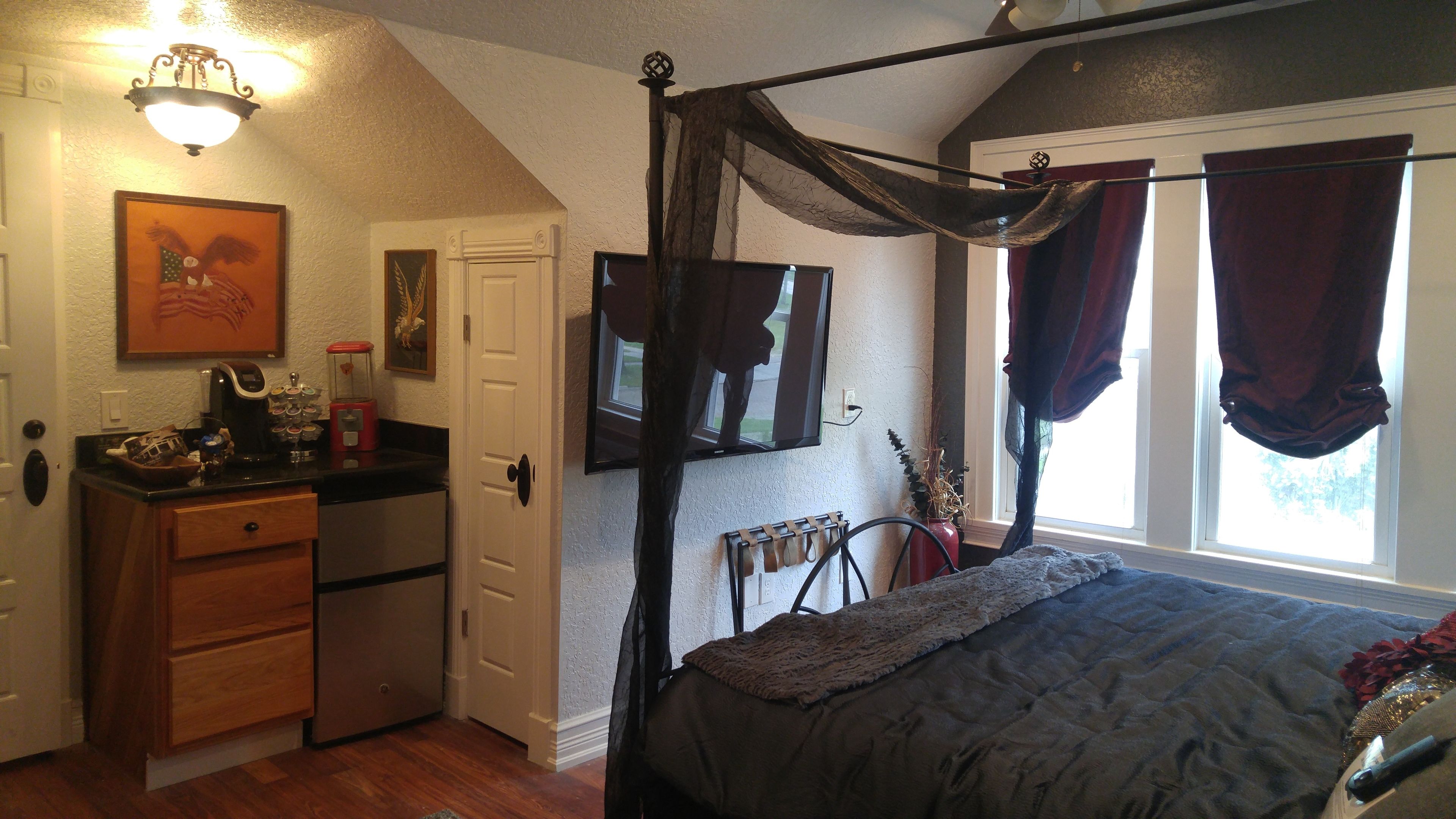 A bedroom with a four-poster bed, a TV mounted on the wall, and a window with curtains.