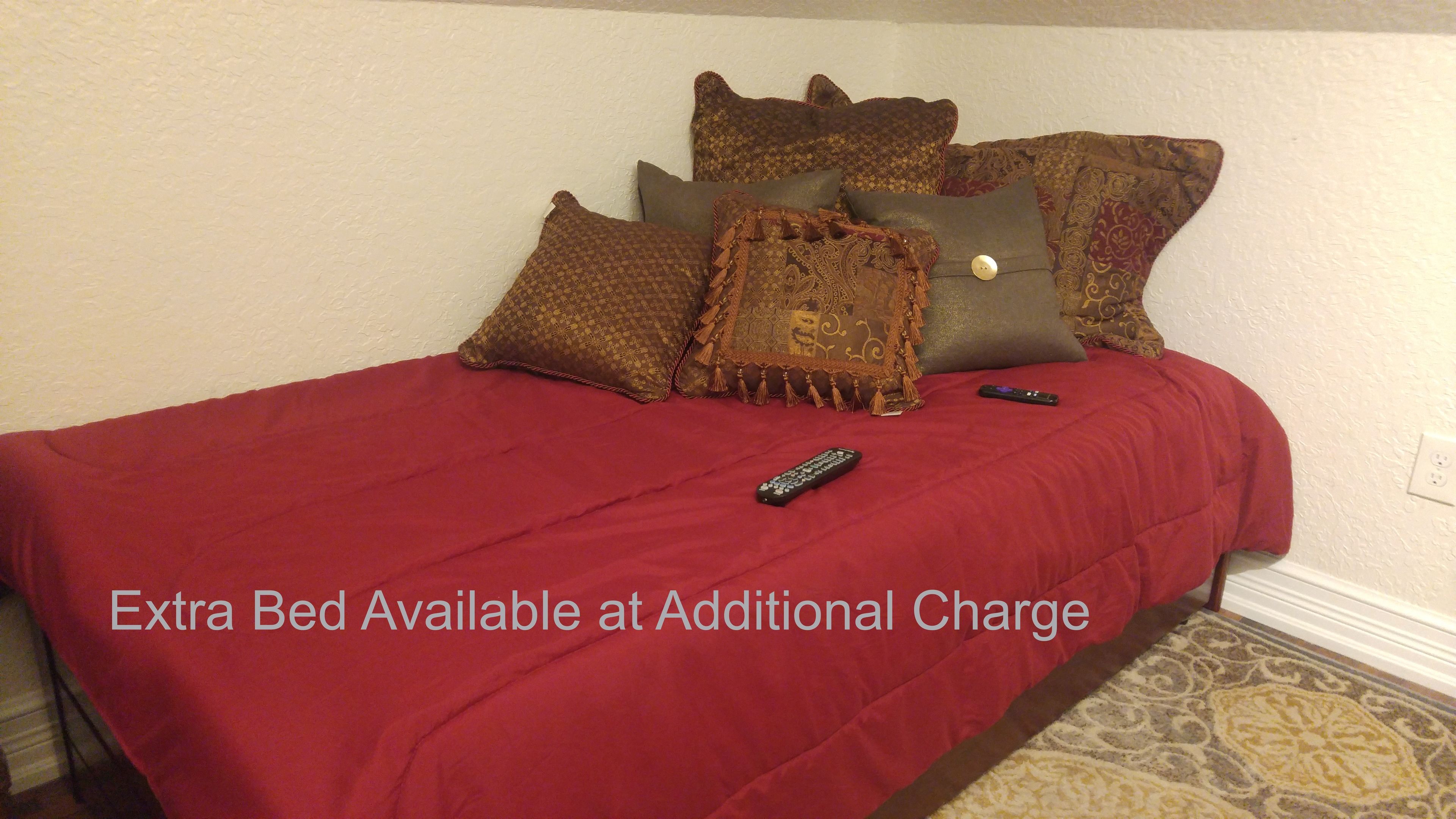 A bed with a red bedspread and multiple pillows, a remote control on the bed, and a text overlay stating "Extra Bed Available at Additional Charge".