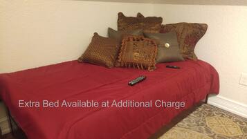 A bed with a red bedspread and multiple pillows, a remote control on the bed, and a text overlay stating "Extra Bed Available at Additional Charge".
