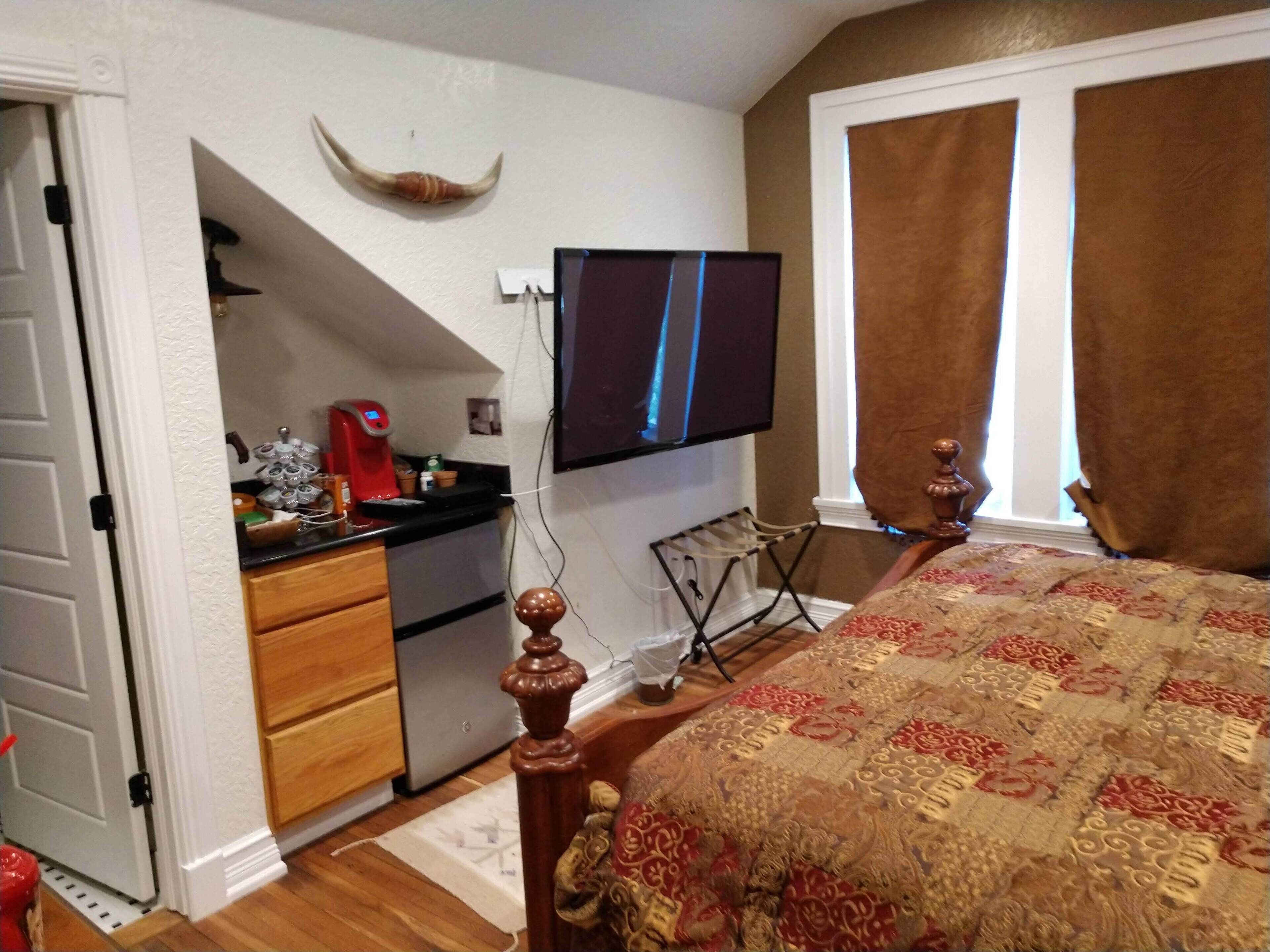 A bedroom with a bed, a desk with a computer, a TV mounted on the wall, and a window with curtains.
