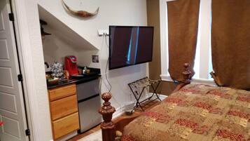 A bedroom with a bed, a desk with a computer, a TV mounted on the wall, and a window with curtains.
