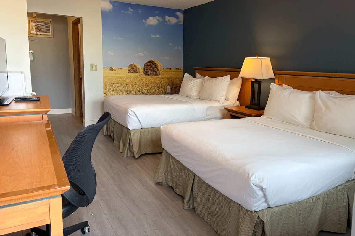 A hotel room with two beds, a desk, a chair, a lamp, and a mural of a field with hay bales.