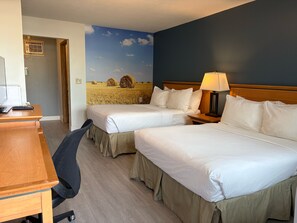 Habitación de hotel con dos camas, un escritorio, una silla, una lámpara y un mural de un campo con pacas de heno.