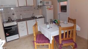 Uma cozinha compacta com armários brancos, fogão, micro-ondas e geladeira. Há uma mesa de jantar com cadeiras e uma janela com cortina verde.