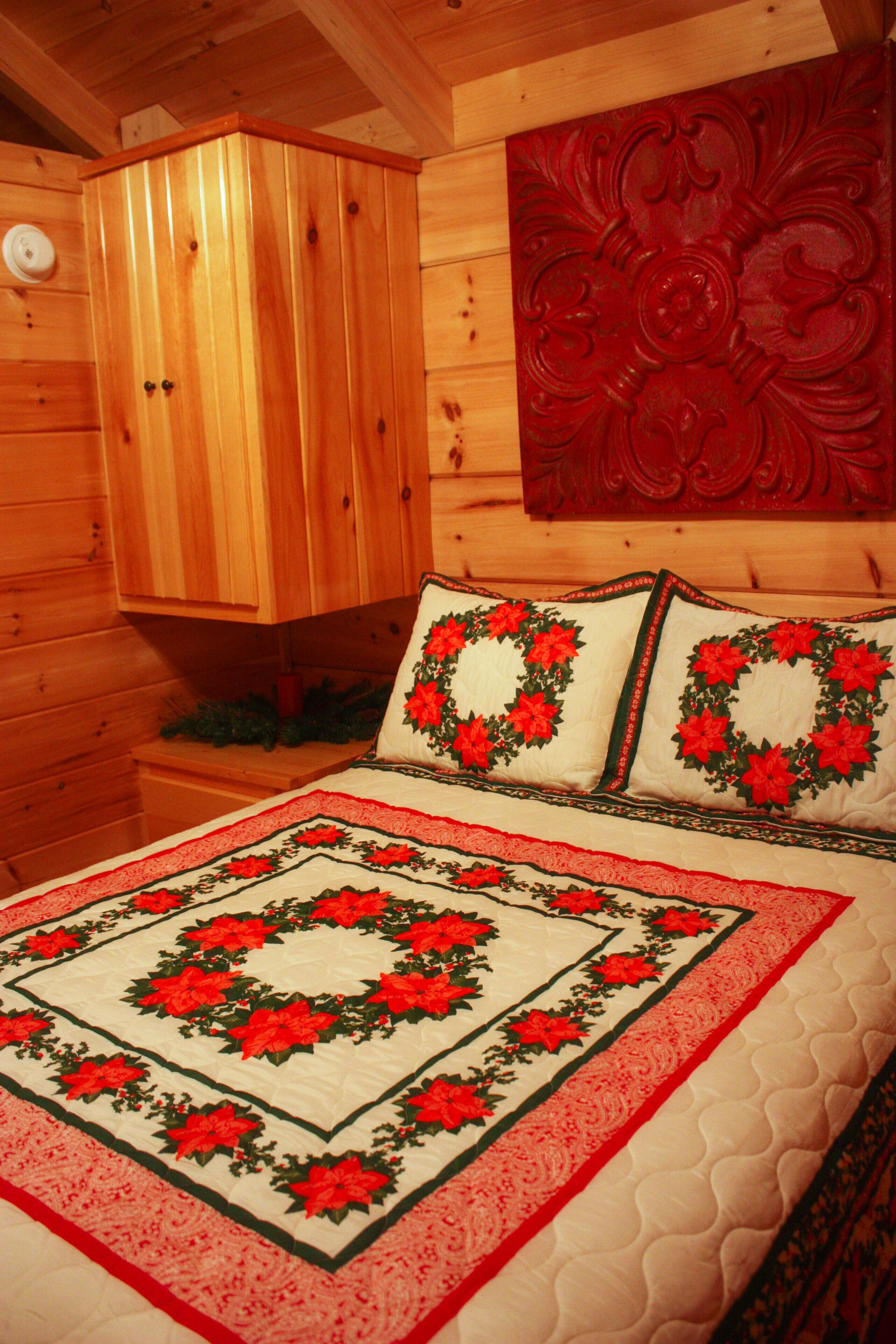 A wooden cabin interior with a bed featuring a red and green floral quilt, a wooden cabinet, and a red decorative panel on the wall.