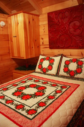 A wooden cabin interior with a bed featuring a red and green floral quilt, a wooden cabinet, and a red decorative panel on the wall.