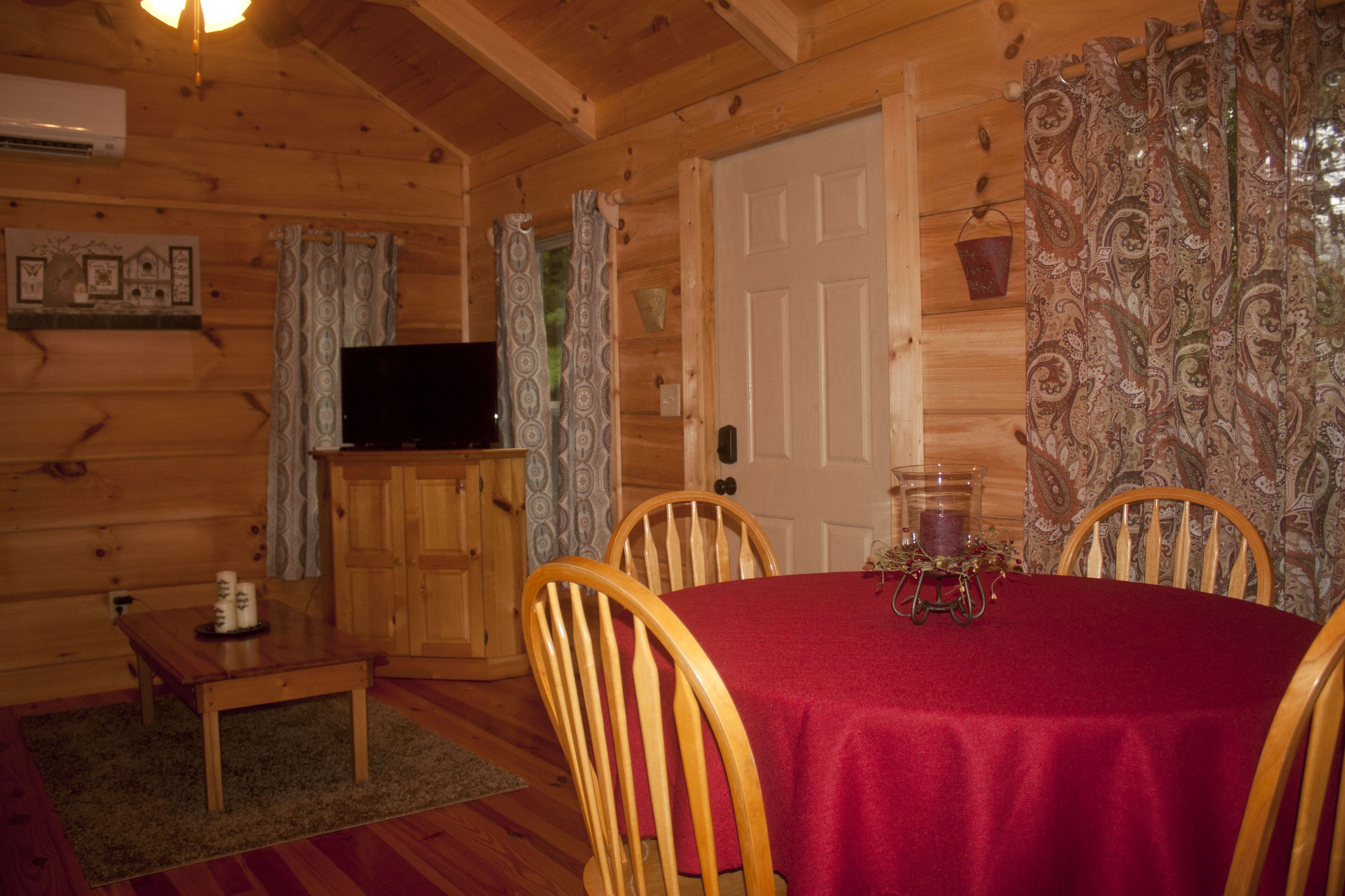 A wooden cabin interior with a dining table, chairs, a television on a wooden cabinet, and patterned curtains.