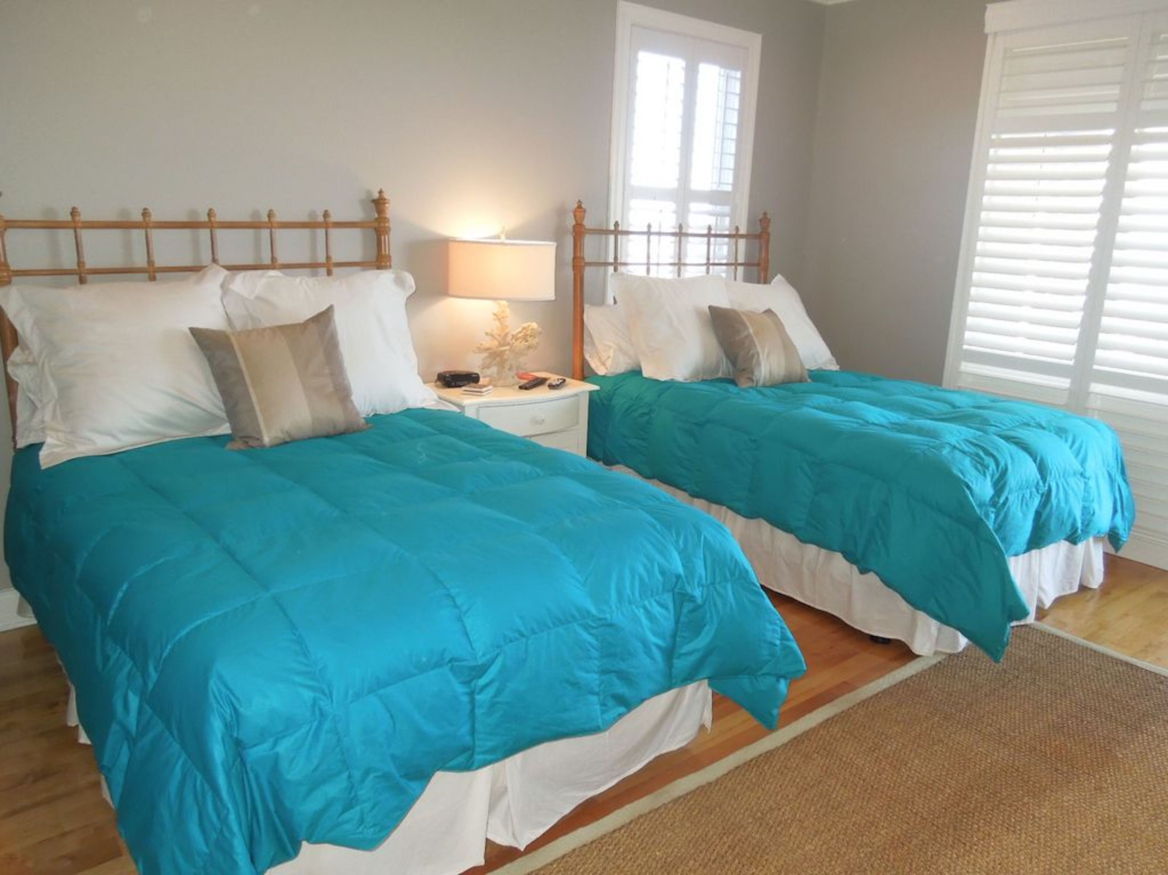 A bedroom with two beds, each with a wooden headboard, white bedding, and a teal duvet. There is a nightstand with a lamp and a small decorative item.