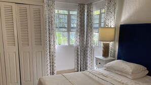 A bedroom with a bed, a nightstand with a lamp, a window with curtains, and a door with louvered panels. - Only 3 mins walk to Beach and Beach Club (holetown)