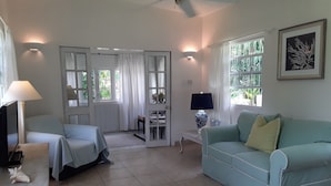 A room with a light blue sofa, a small table with a lamp, a television, and a door leading to an outdoor area. - Only 3 mins walk to Beach and Beach Club (holetown)