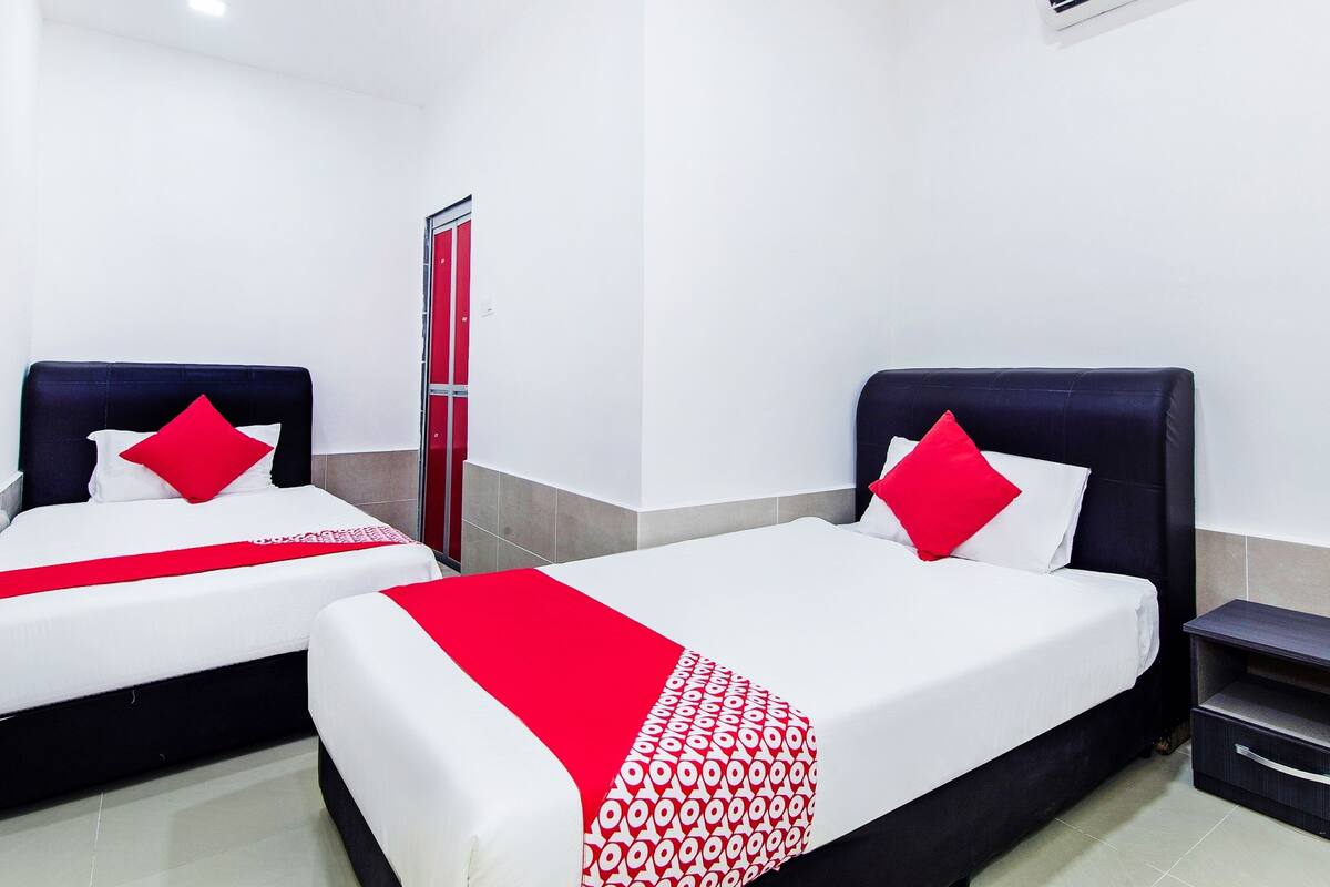 Ein Hotelzimmer mit zwei Betten, jeweils mit einem roten Kissen und einer weiß-roten Bettdecke. Ein kleiner Nachttisch mit Schublade ist vorhanden, sowie eine Wandaircondition.