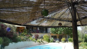 En pool under et stråtækt tag, omgivet af frodig vegetation og en bygning i baggrunden.