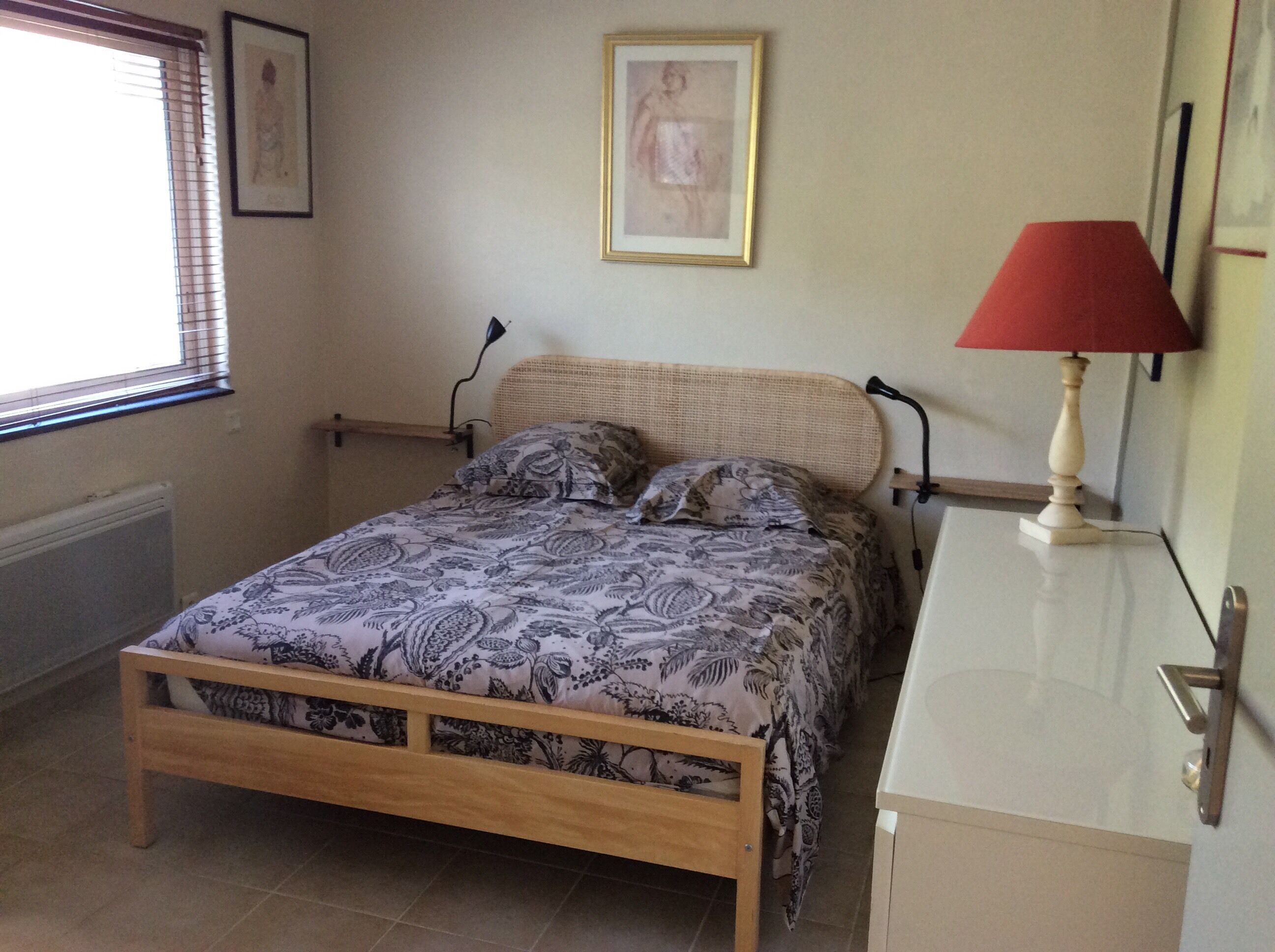 A bedroom with a wooden bed, a bedside table with a lamp, a radiator, and a window with blinds.