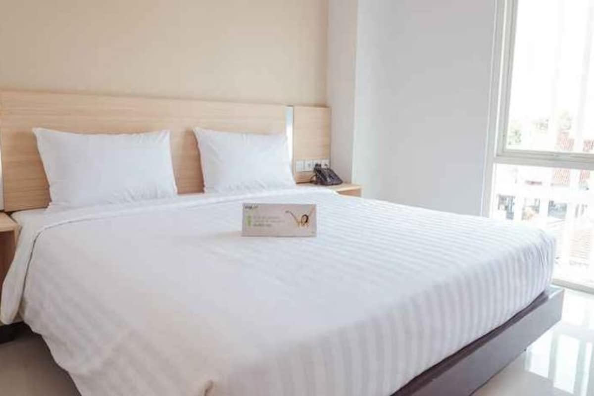 Quarto de hotel com uma cama grande, cabeceira de madeira, mesa de cabeceira com telefone e janela com vista.