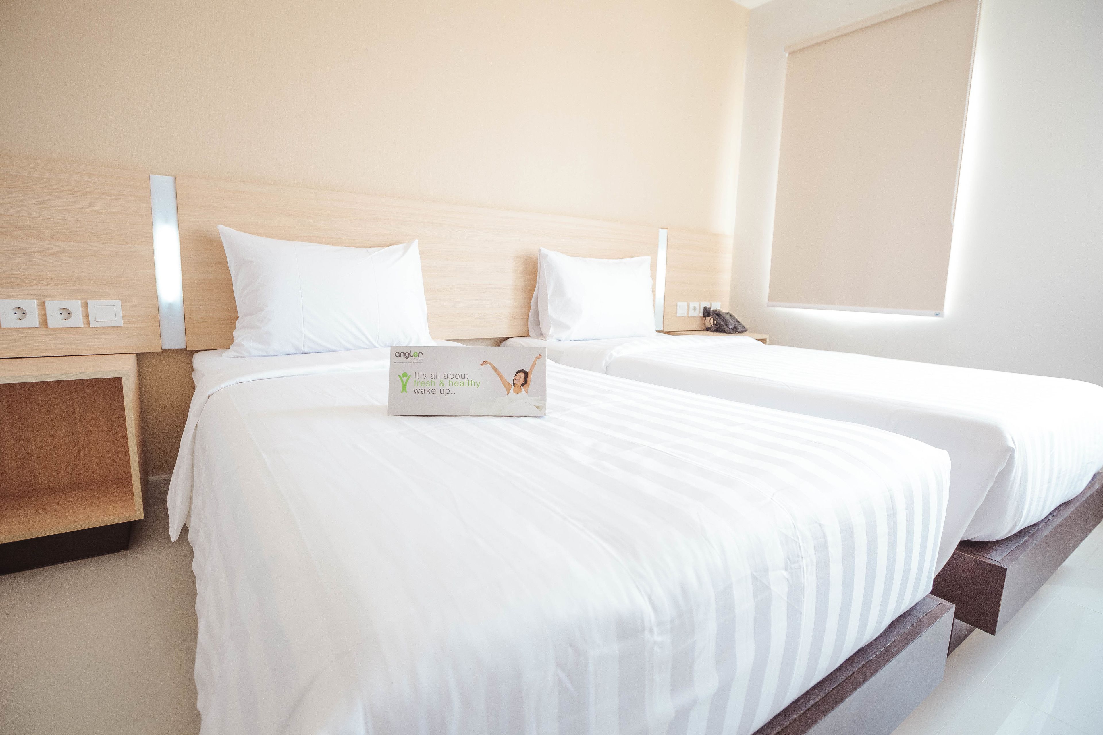 A hotel room with two beds, wooden headboards, white linens, a bedside table with a lamp, and a window with blinds.