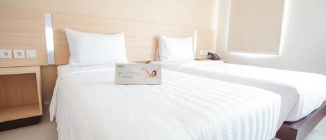 A hotel room with two beds, wooden headboards, white linens, a bedside table with a lamp, and a window with blinds.