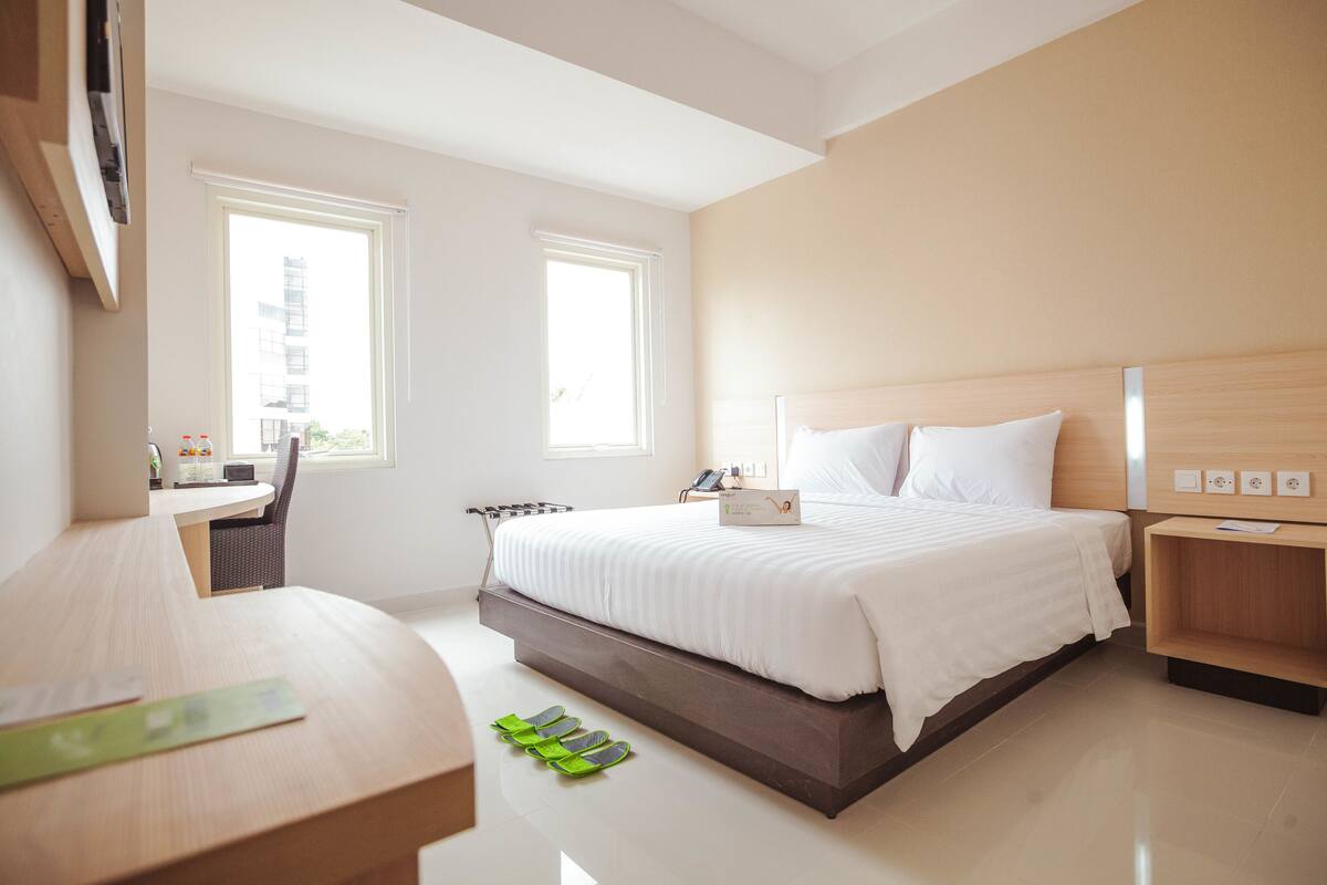 Habitación de hotel moderna con una cama grande, un escritorio y una silla. Dos ventanas ofrecen luz natural.