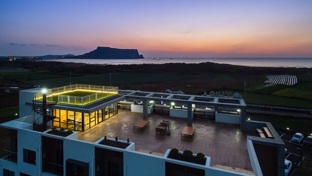Una terraza en la azotea con cerramiento de vidrio, mobiliario exterior y vistas al mar y colinas a lo lejos.