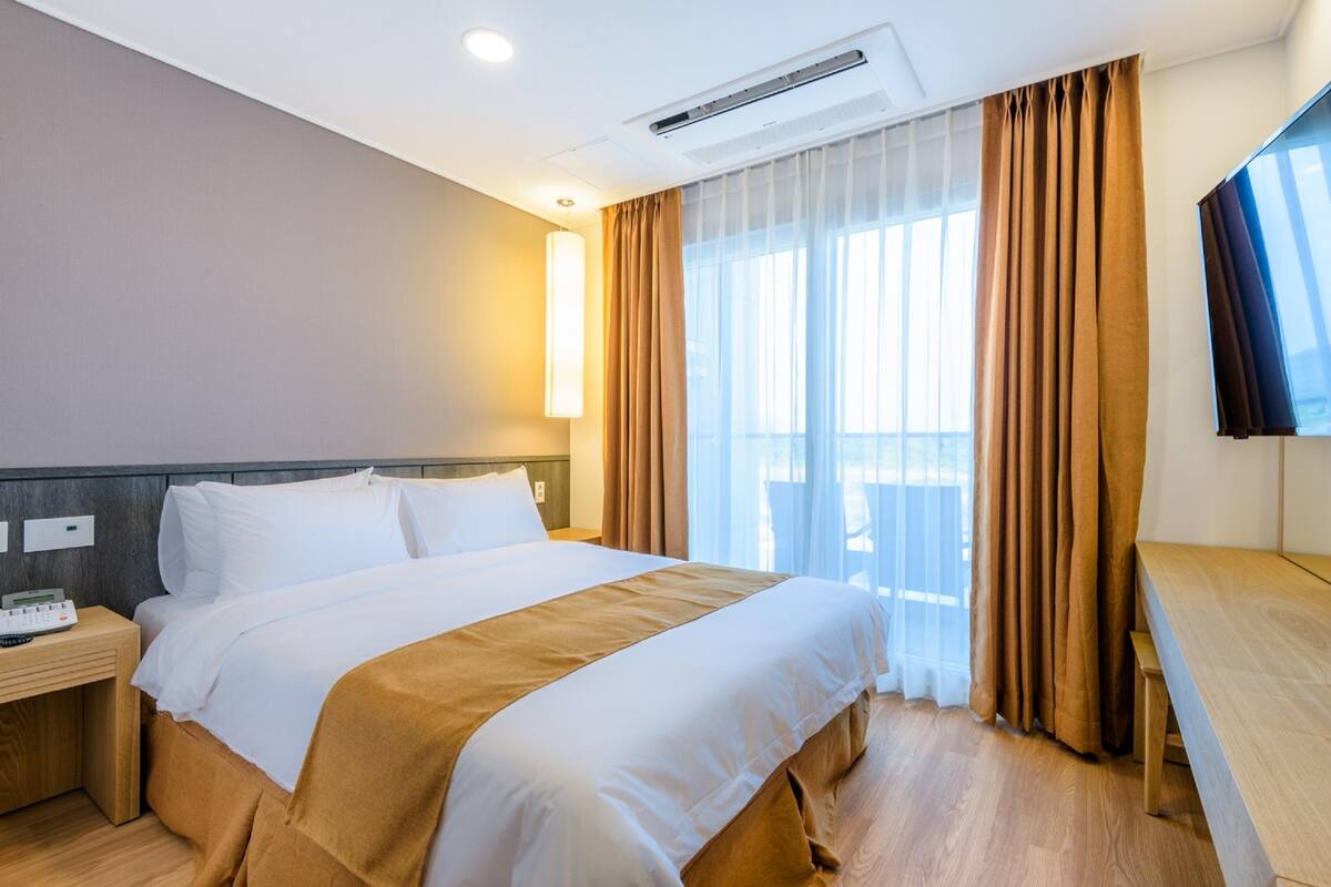 A hotel room with a large bed, a bedside table with a phone, a desk with a chair, and a large window with curtains.