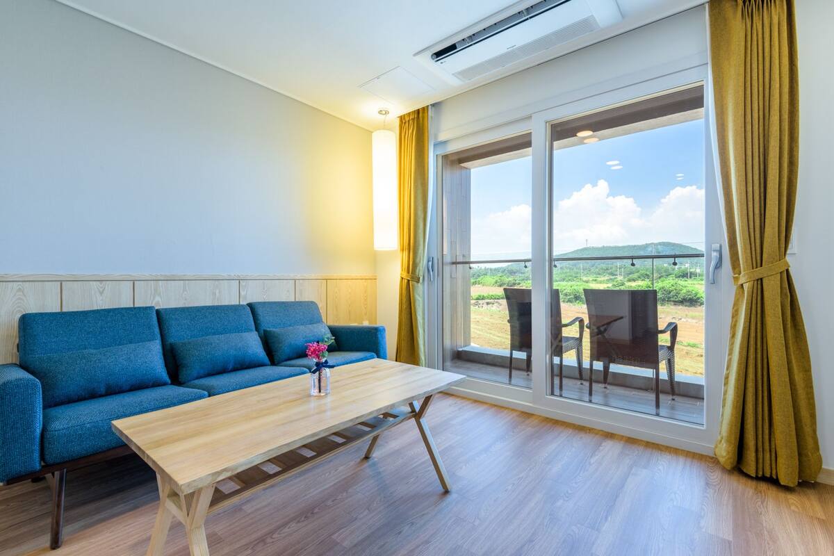 A room with a blue sofa, a wooden table, and a view of the outdoors through large windows.