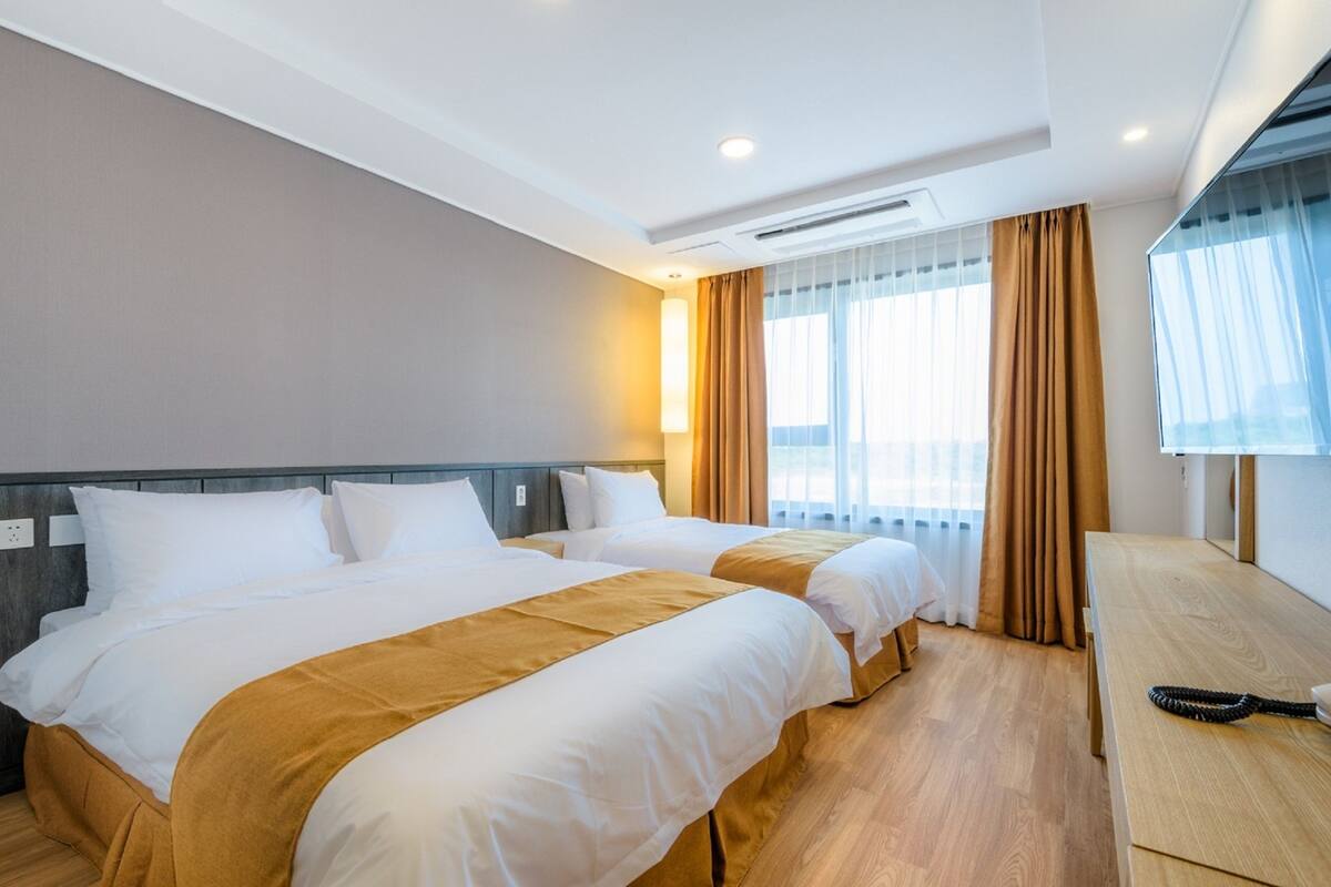 A hotel room with two beds, a wooden desk, and a large window with curtains.